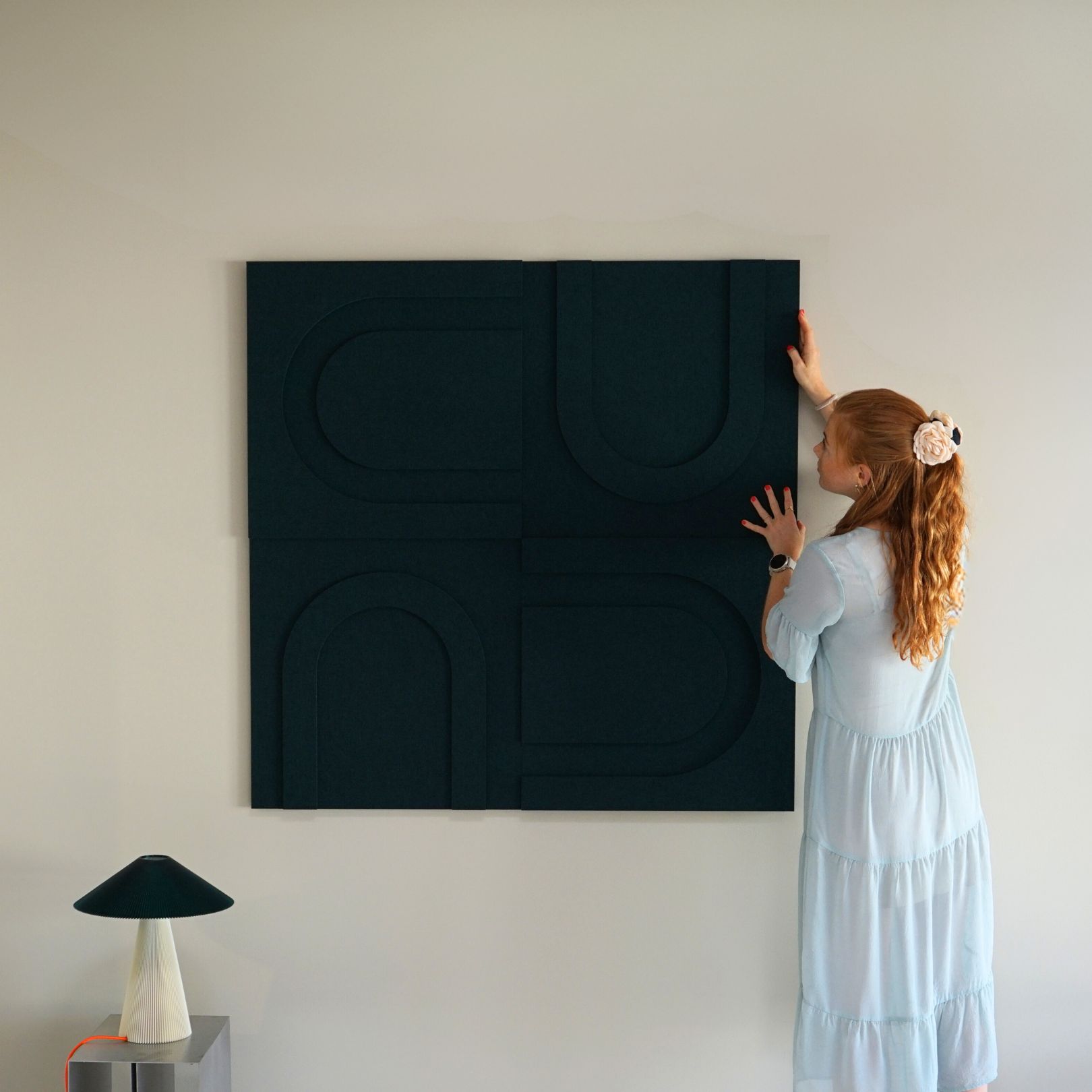 Woman installing dark green geometric acoustic art panel from Arturel, designed for sound absorption and modern interior aesthetics.